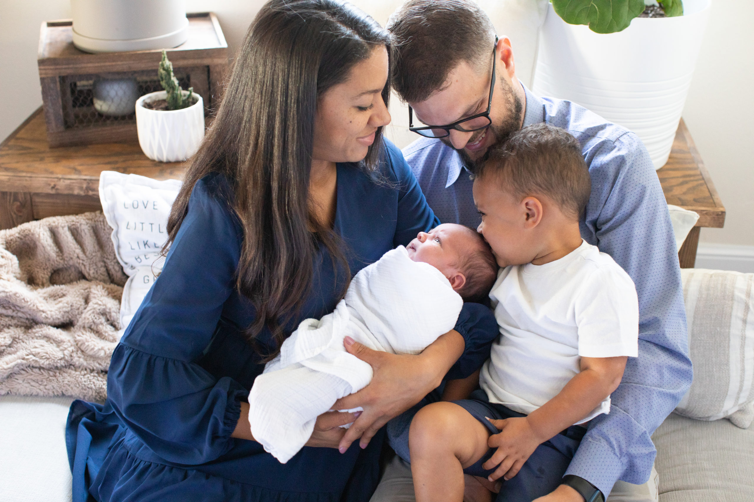 Mom in navy blue dress holds newborn baby boy, while dad in light blue shirt holds older son. They are all cuddled together on cushions